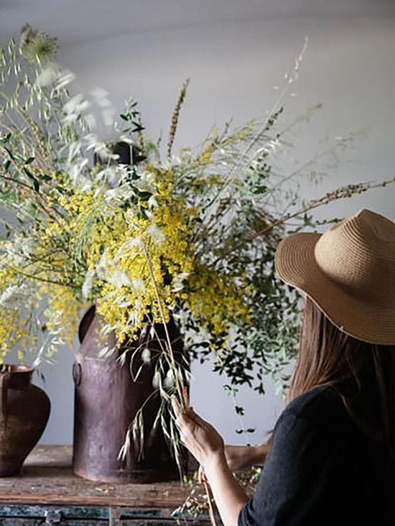 A woman in a straw hat arranging mimosa and olive branches in a clay urn